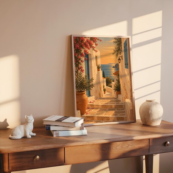 Mediterranean coastal village wall art print leaning on a wooden desk against a warm beige wall in soft window sunlight.