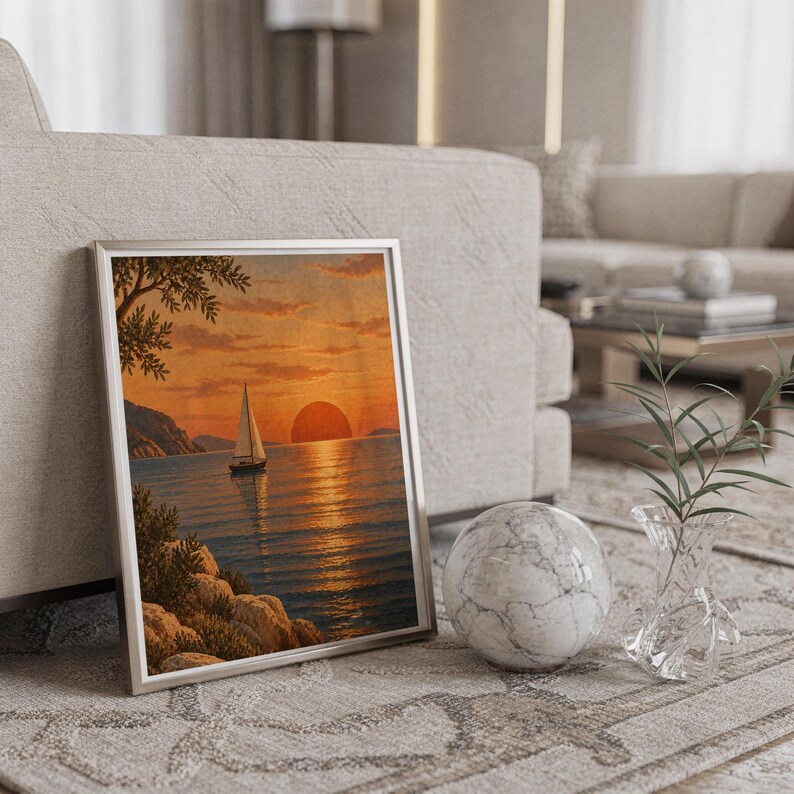 Sunset sailboat wall art print in a silver frame leaning against a sofa beside a decorative marble sphere.