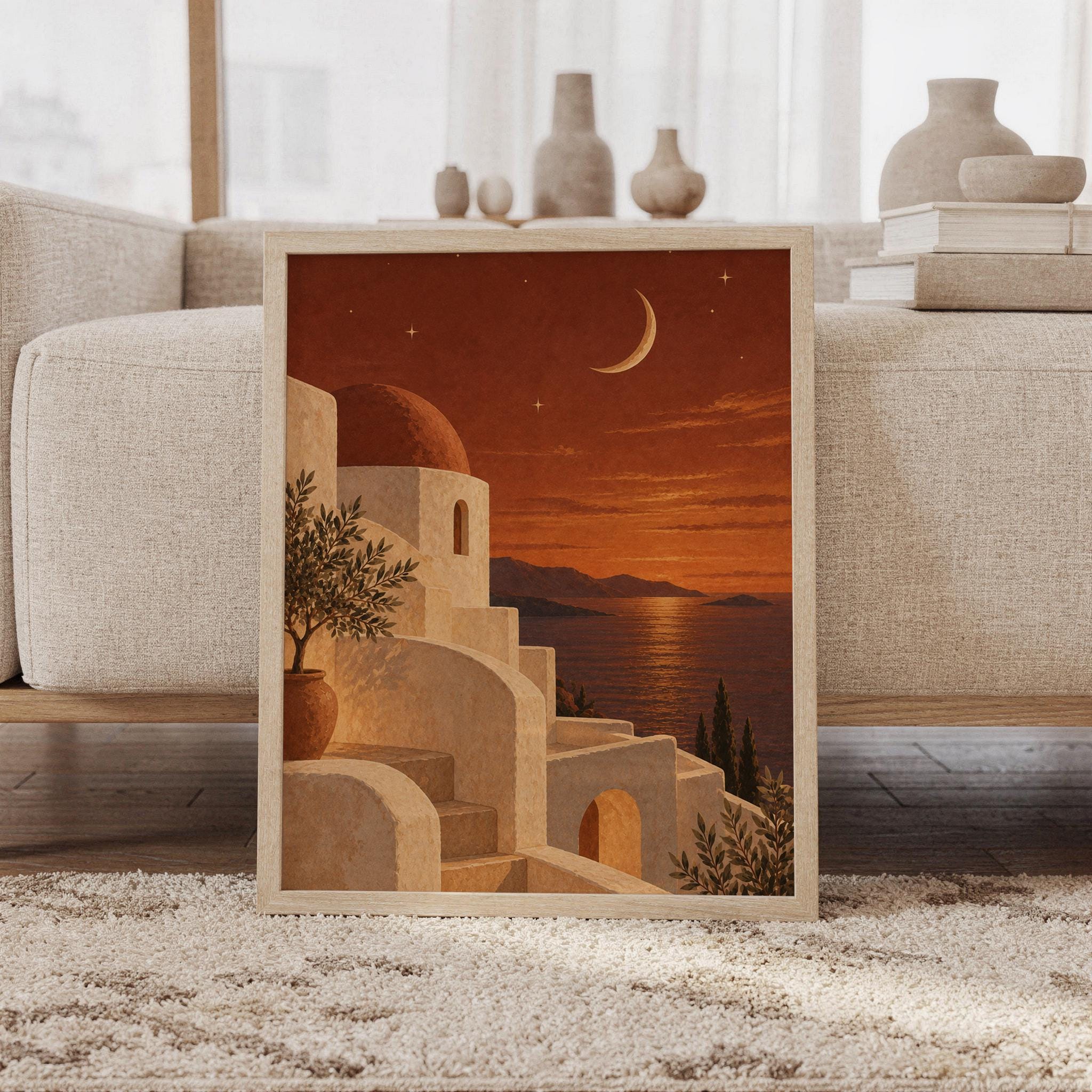 Mediterranean moonlit coastal wall art print featuring warm terracotta skies, creamy stone architecture, a glowing crescent moon, olive trees, and a tranquil sea view, framed and leaning against a beige sofa on a textured rug in a bright Scandinavian living room.