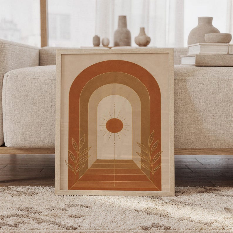 Boho arch sun wall art print featuring terracotta, warm tan, caramel brown, and cream tones with layered geometric arches, a radiant sun, and delicate botanical leaves, framed and leaning against a beige sofa on a textured rug in a bright Scandinavian living room.