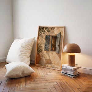 Mediterranean blue shutter wall art print leaning on a herringbone wood floor beside cream cushions and a wooden mushroom lamp.
