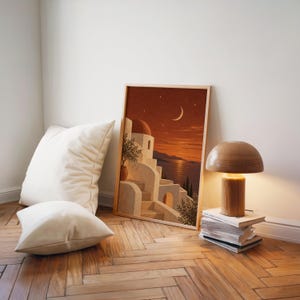 Mediterranean moonlit coastal wall art print leaning on a herringbone wood floor beside cream cushions and a wooden mushroom lamp.