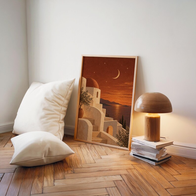 Mediterranean moonlit coastal wall art print leaning on a herringbone wood floor beside cream cushions and a wooden mushroom lamp.