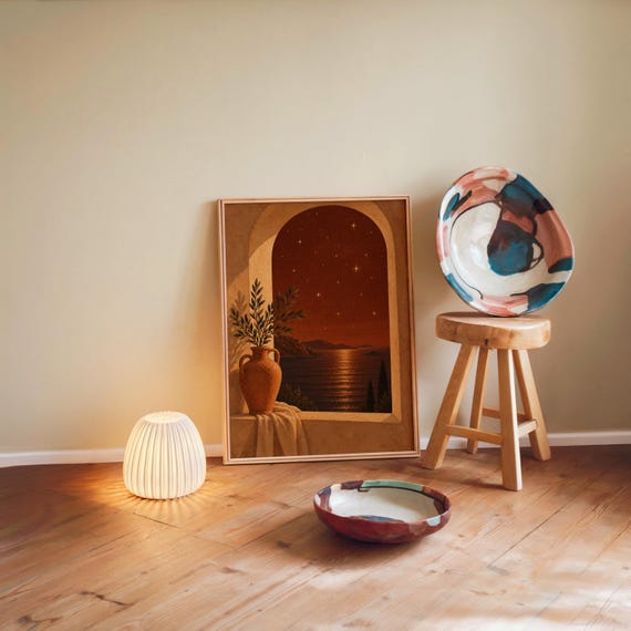 Mediterranean night sea wall art print leaning against a warm beige wall on a wooden floor beside a stool and a glowing lamp.