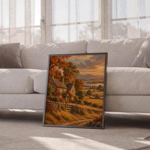 Autumn cottage landscape wall art print leaning against a white sofa in a bright living room with sheer curtains.
