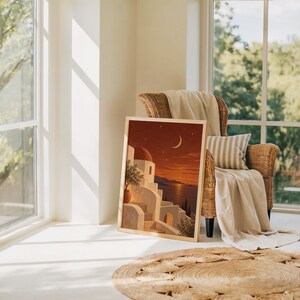 Mediterranean moonlit coastal wall art print leaning on the floor beside a rattan armchair and jute rug in a bright sunlit room.