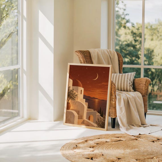 Mediterranean moonlit coastal wall art print leaning on the floor beside a rattan armchair and jute rug in a bright sunlit room.