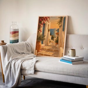 Mediterranean coastal village wall art print leaning on a cream sofa against a beige wall beside books and a mug.