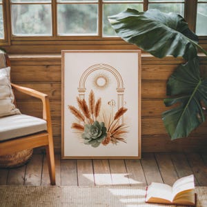 Celestial sun and pampas grass botanical poster framed on dark hardwood flooring near a sunlit window with large green monstera plant leaves, warm sunlight casting shadows on the wooden floor and wall in a cozy modern bohemian living room environment.