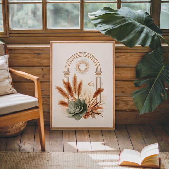Celestial sun and pampas grass botanical poster framed on dark hardwood flooring near a sunlit window with large green monstera plant leaves, warm sunlight casting shadows on the wooden floor and wall in a cozy modern bohemian living room environment.