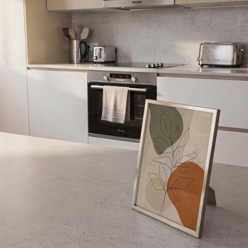 Small framed botanical line art piece leaning against the backsplash on a modern kitchen counter.
