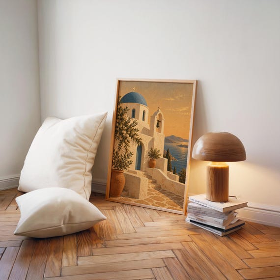 Santorini church wall art print leaning on a herringbone wood floor beside cream cushions and a wooden mushroom lamp.