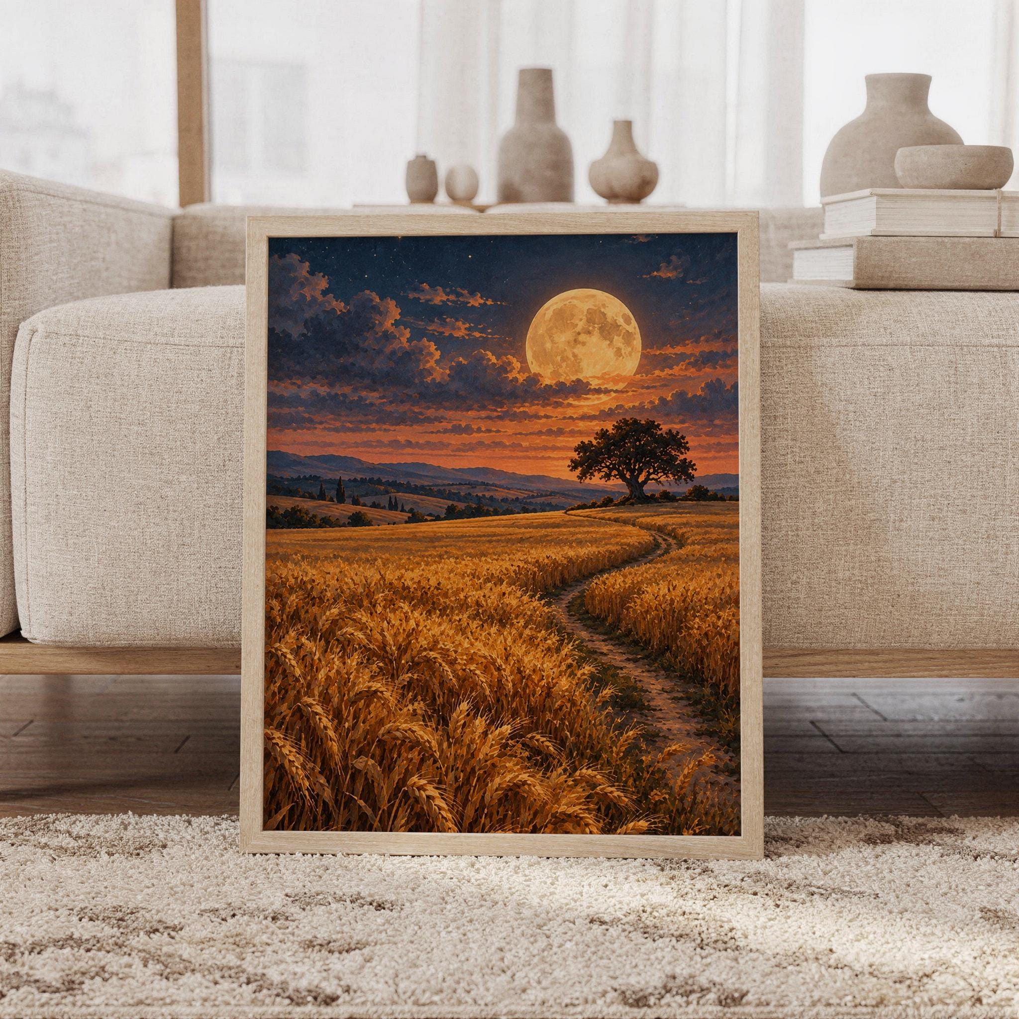 Moon wall art print featuring glowing amber wheat, a golden full moon, dusky navy skies, copper clouds, rolling hills, and a lone tree in a painterly countryside style, framed and leaning against a beige sofa on a textured rug in a bright Scandinavian living room.
