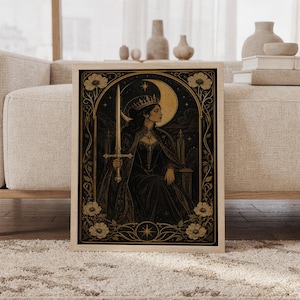 Gothic moon queen wall art print featuring antique gold, black, and warm sepia tones, a crowned warrior queen holding a sword before a glowing crescent moon, ornate florals, and stars, framed and leaning against a beige sofa on a textured rug in a bright Scandinavian living room.