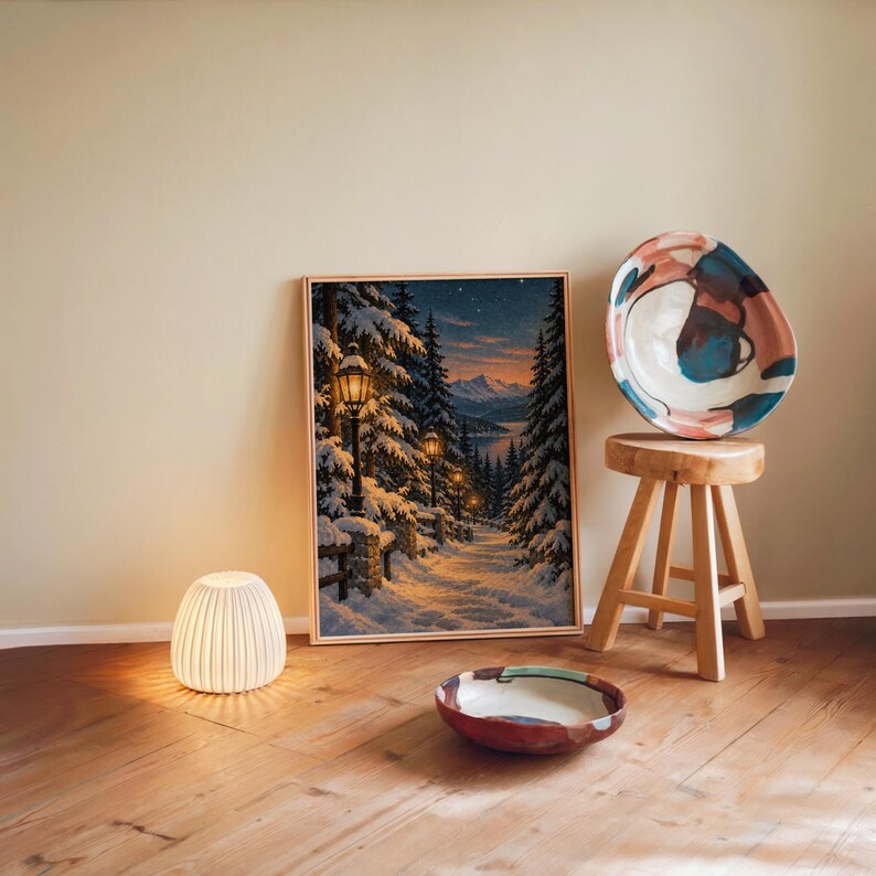 Snowy forest path wall art print leaning against a warm beige wall on a wooden floor beside a stool and a glowing lamp.