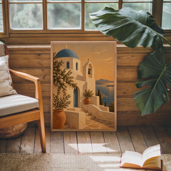 Santorini church wall art print leaning against warm wood paneling beside a wooden armchair and a monstera plant.