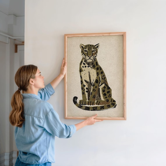 A woman in a denim shirt holds a framed print of a sitting leopard with dark green and brown spots on a light background, against a light wall.