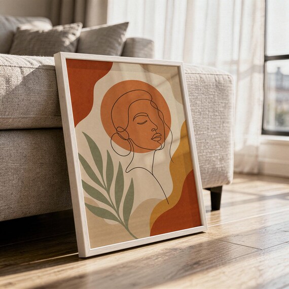 Abstract line art face print in a white frame leaning against a gray sofa on a sunlit wooden floor.