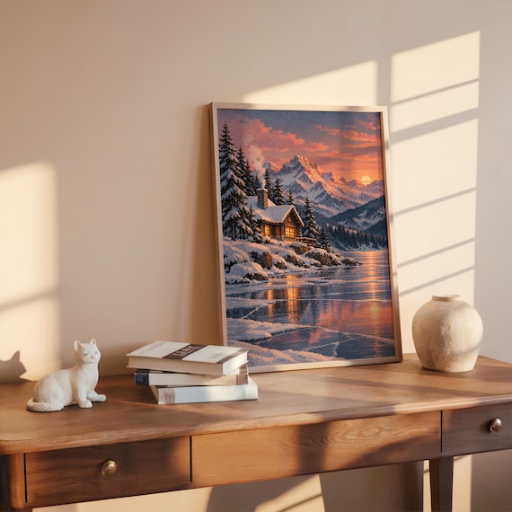 Winter cabin mountain wall art print leaning on a wooden desk against a warm beige wall in soft window sunlight.