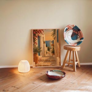 Mediterranean coastal village wall art print leaning against a warm beige wall on a wooden floor beside a stool and a glowing lamp.