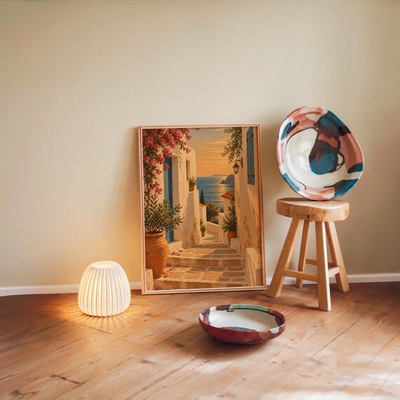 Mediterranean coastal village wall art print leaning against a warm beige wall on a wooden floor beside a stool and a glowing lamp.