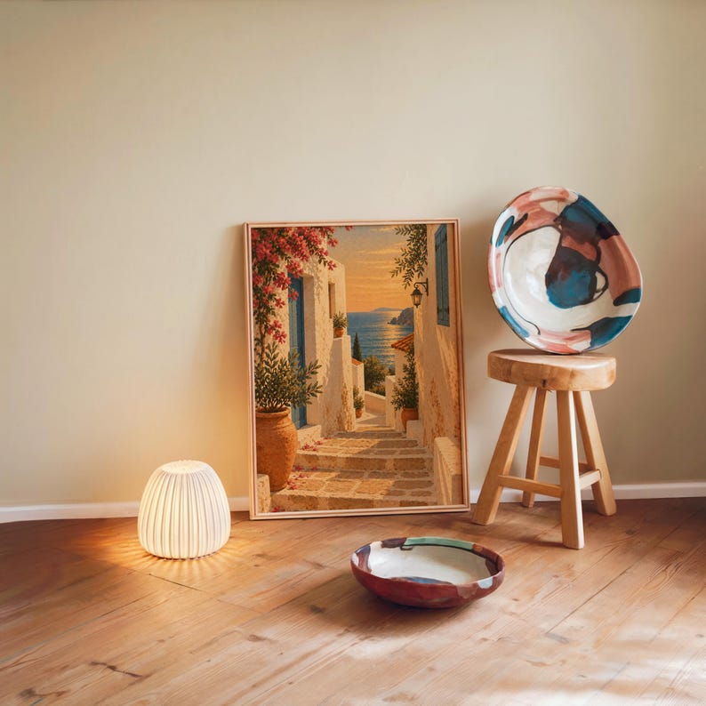 Mediterranean coastal village wall art print leaning against a warm beige wall on a wooden floor beside a stool and a glowing lamp.