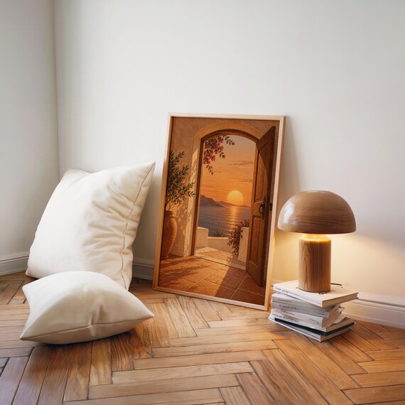 Mediterranean sunset doorway wall art print leaning on a herringbone wood floor beside cream cushions and a wooden mushroom lamp.