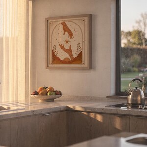 Earthy terracotta hands tarot print framed on kitchen wall illuminated by warm sunset light.