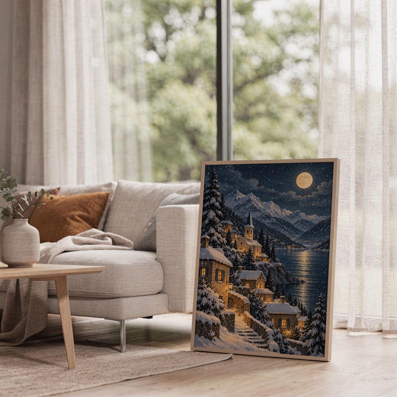 Snowy mountain village wall art print leaning on the floor beside a cream armchair next to large garden windows.