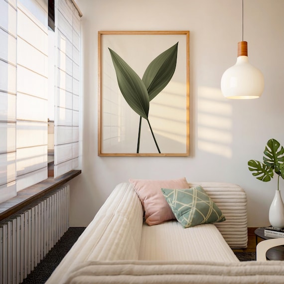 A framed print of two dark green leaves on a light background hangs above a white sofa with pink and green pillows, in a room with a large window.