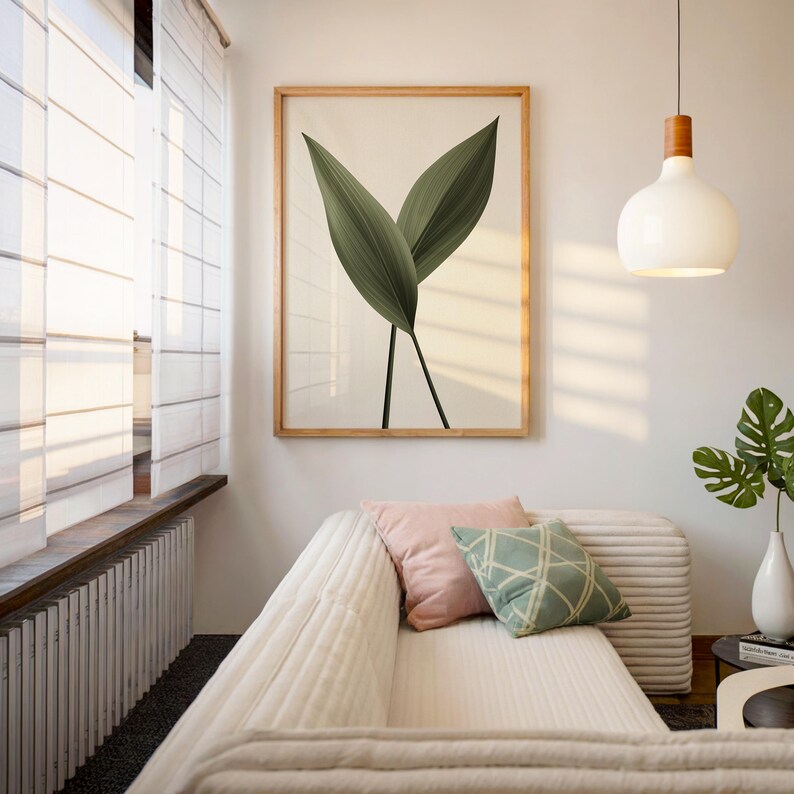 A framed print of two dark green leaves on a light background hangs above a white sofa with pink and green pillows, in a room with a large window.