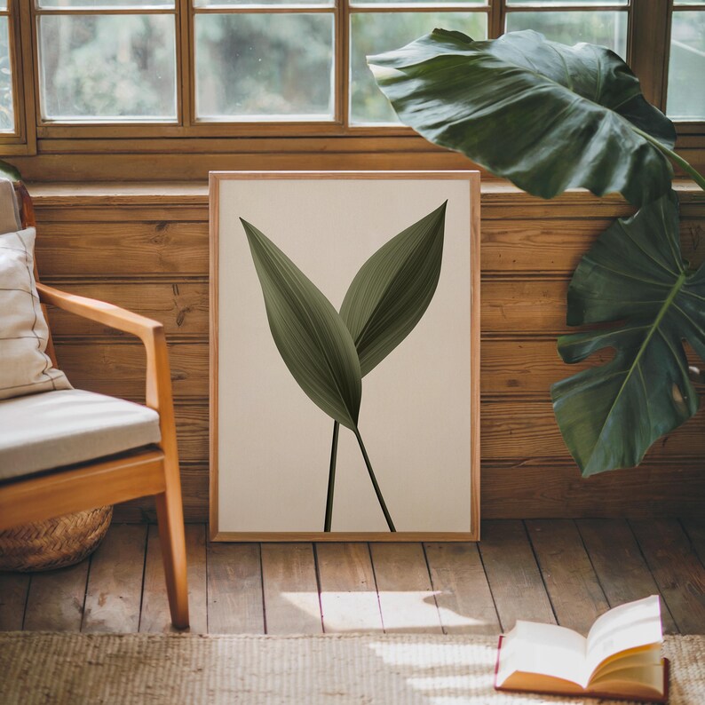 A framed print of two dark green leaves on a light background sits on a wooden floor, next to a wooden chair and a large plant, with an open book nearby.