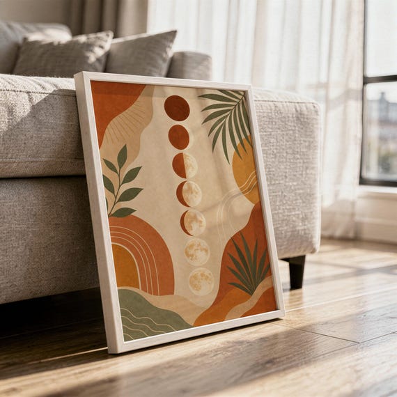 Boho moon phases art print in a white frame leaning against a gray sofa on a sunlit wooden floor.