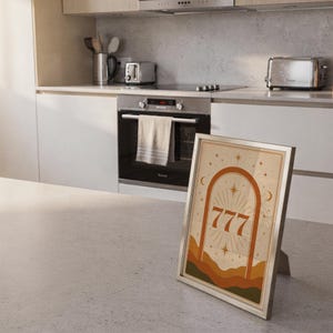 Framed terracotta 777 celestial art print resting on the floor in a bright, modern kitchen interior.