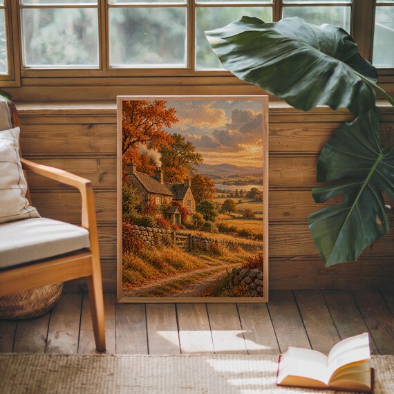 Autumn cottage landscape wall art print leaning against warm wood paneling beside a wooden armchair and a monstera plant.