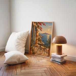 Mediterranean balcony wall art print leaning on a herringbone wood floor beside cream cushions and a wooden mushroom lamp.