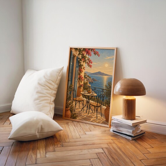 Mediterranean balcony wall art print leaning on a herringbone wood floor beside cream cushions and a wooden mushroom lamp.