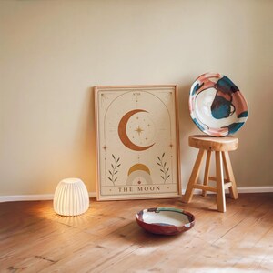 Aesthetic neutral moon tarot card print framed on wooden floor with ambient night lamp.