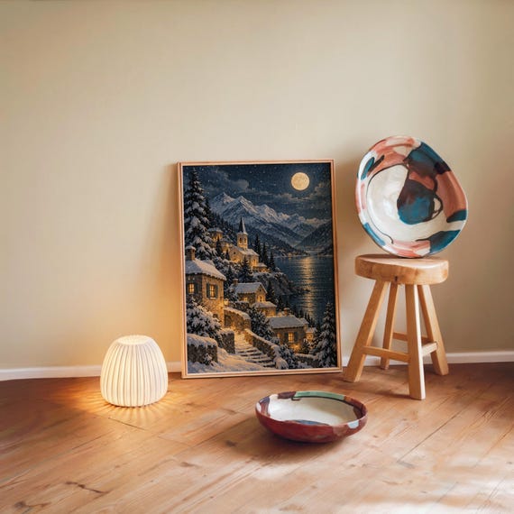 Snowy mountain village wall art print leaning against a warm beige wall on a wooden floor beside a stool and a glowing lamp.
