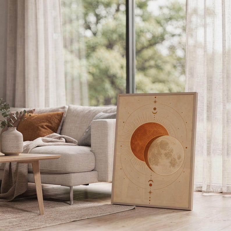 Sun and moon celestial wall art print leaning on the floor beside a cream armchair next to large garden windows.