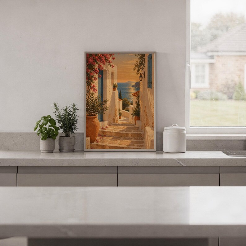 Mediterranean coastal village wall art print standing on a white kitchen counter beside potted herbs.
