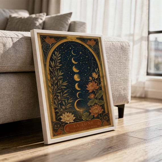 Moon phase wall art print in a white frame leaning against a gray sofa on a sunlit wooden floor.