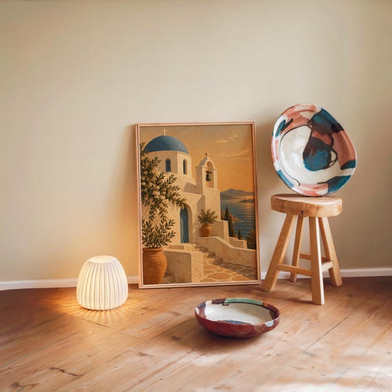 Santorini church wall art print leaning against a warm beige wall on a wooden floor beside a stool and a glowing lamp.