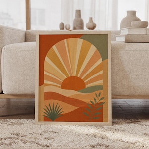 Boho sun arch wall art print of a rising sun inside an arch above layered hills with leaves in terracotta, ochre, peach and muted sage, framed and leaning against a beige sofa on a textured rug in a bright Scandinavian living room.