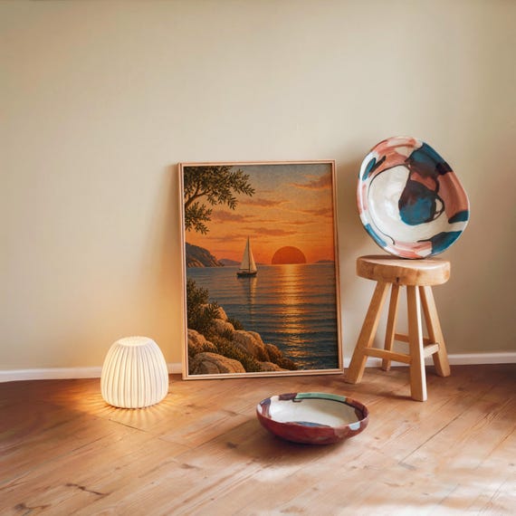 Sunset sailboat wall art print leaning against a warm beige wall on a wooden floor beside a stool and a glowing lamp.