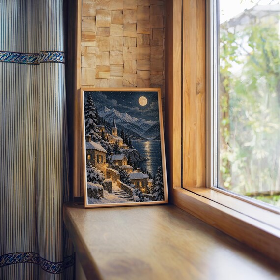 Snowy mountain village wall art print standing on a wooden window sill in warm natural light.