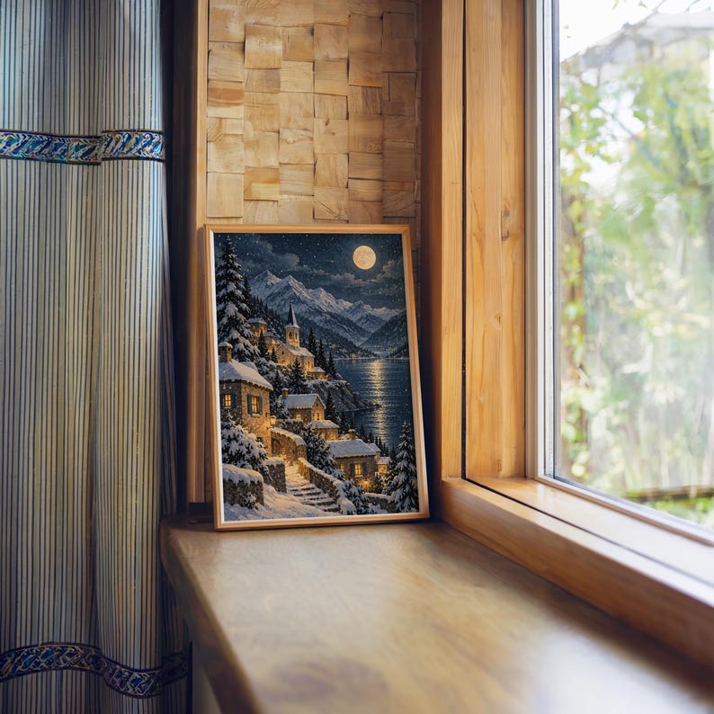 Snowy mountain village wall art print standing on a wooden window sill in warm natural light.