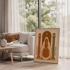 Framed abstract terracotta print leaning against a sofa next to a low wooden coffee table in a bright room.