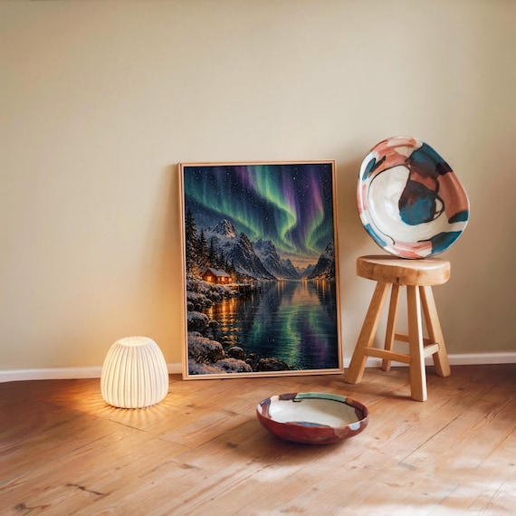 Aurora borealis mountain landscape wall art print leaning against a warm beige wall on a wooden floor beside a stool and a glowing lamp.