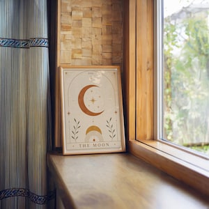 Single framed The Moon art print resting on a wooden windowsill next to a window.